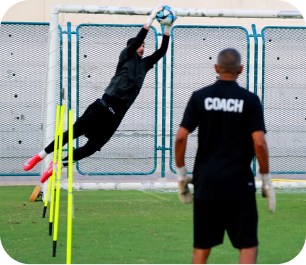 Goalkeeper leaping to make a high save during training while a coach in a "COACH" shirt watches
