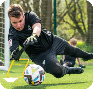 Goalkeeper in all-black kit diving low to make a save during training on artificial turf