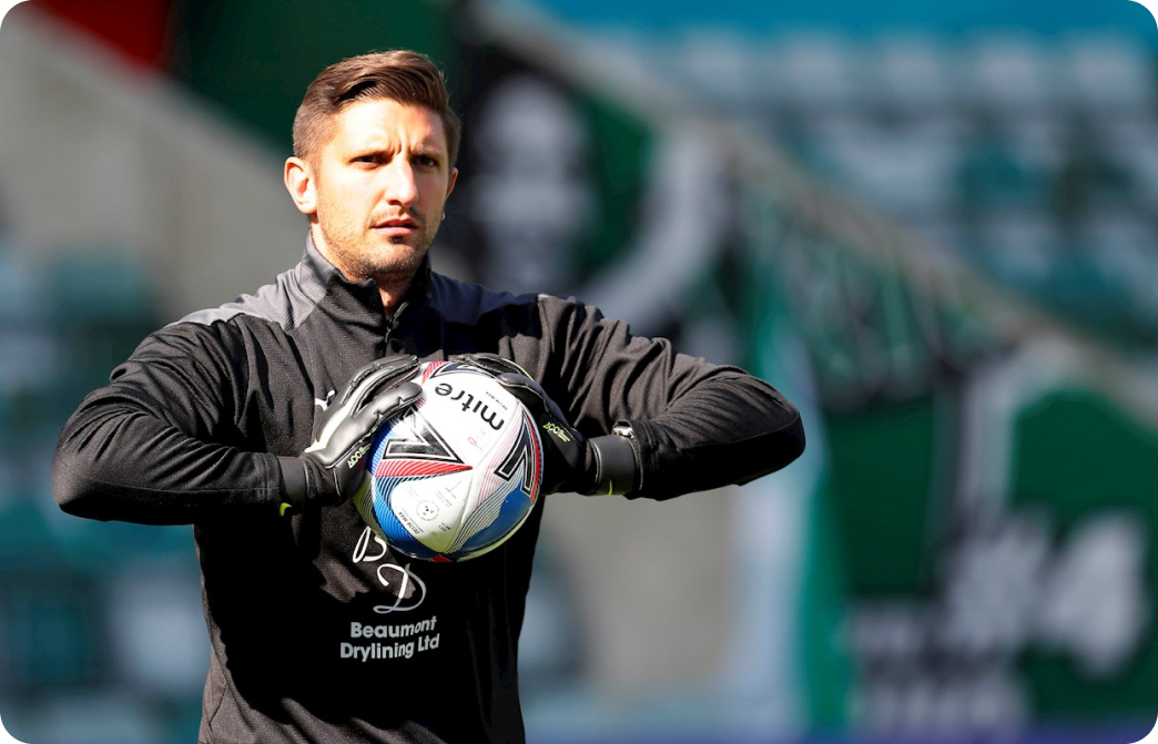 Goalkeeper in black kit and gloves holding a Mitre ball during warm-up at a stadium