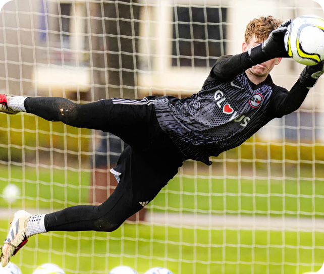 Goalkeeper diving midair to catch a soccer ball in front of the goal net