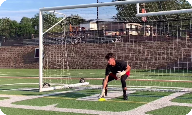 Goalkeeper in black kit in a low ready stance during solo training on an artificial turf field