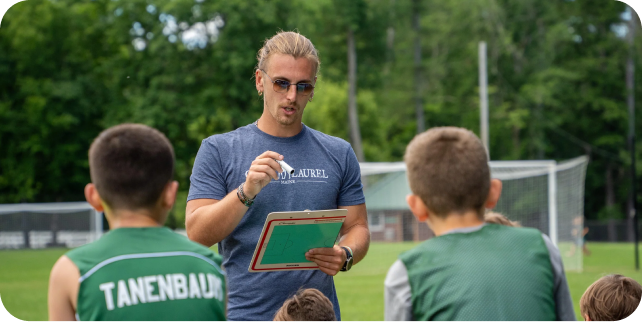 A coach with a clipboard giving instructions to young soccer players in green kits on an outdoor field