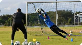 A goalkeeper in blue dives to make a save during an outdoor training session with a coach watching